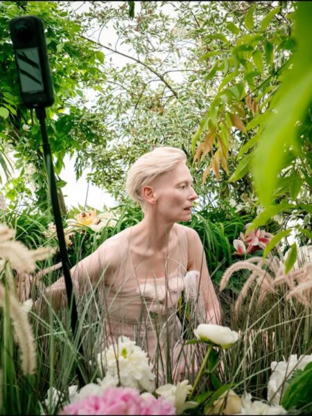 Tilda Swinton w pastelowej sukni, siedząca wśród gęstej roślinności, otoczona kwiatami w odcieniach bieli, różu i zieleni, z naturalnym światłem wpadającym przez liście, podczas realizacji zdjęcia w plenerowej scenografii.