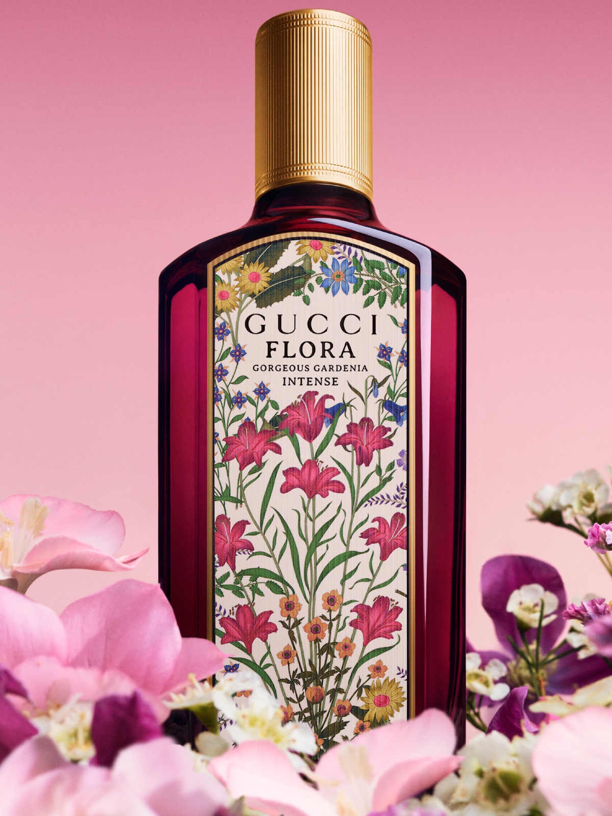 Gucci Flora Gorgeous Gardenia Eau de Parfum Intense: Produktowe zdjęcie flakonu Gucci Flora Gorgeous Gardenia Intense ustawionego wśród pastelowych kwiatów na tle łagodnego gradientu w odcieniach różu. Kompozycja wizualna emanuje romantyzmem i subtelnym luksusem, idealnie oddając kwiatowo-drzewny charakter zapachu. Flakon centralnie wyeksponowany z pełnym widokiem na zdobioną etykietę.