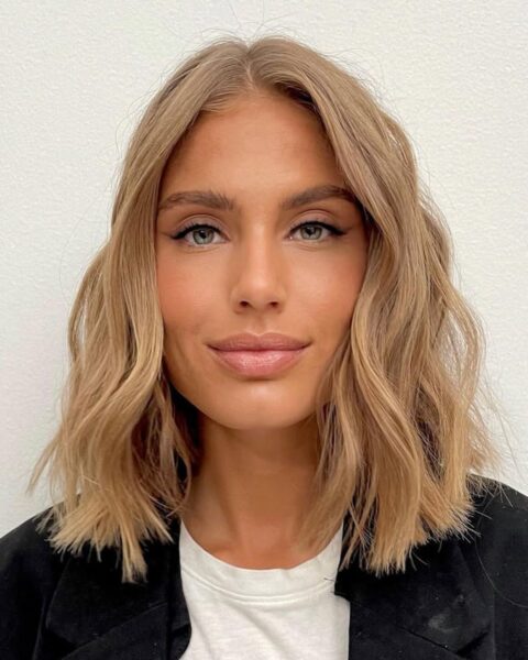 Zdjęcie kobiety ze zbliżeniem na jej twarz. Ma makijaż oczu z eyelinerem na powiece. Ma blond włos w stylu long boba. Ubrana jest w biały top i czarną marynarkę.