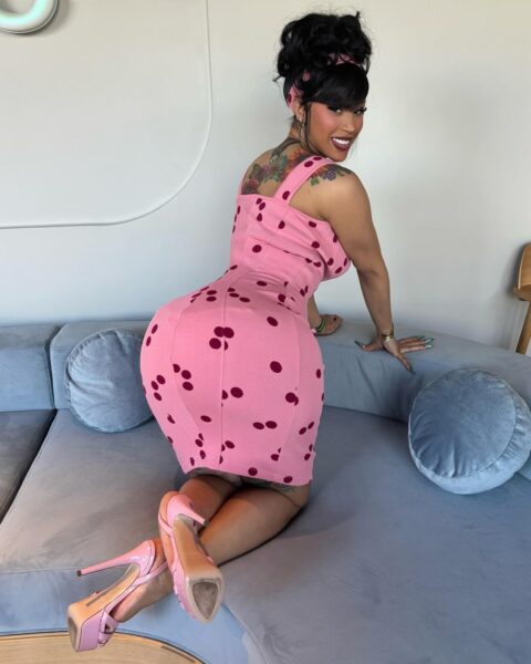 OnlyFans celebryci: na zdjęciu jest Cardi Bi w różowej sukience w czerwone groszki. Ma włosy spięte w kok. Buty wysokie, różowe, na obcasie. Pozuje klęcząc na kanapie i wypina tyłek.