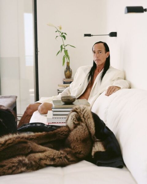 Na zdjęciu jest projektant Rick Owens, który pozuje na OnlyFans. Gwiazdor jest ubrany w białą marynarkę oversize. Ma ciemne włosy rozpuszczone na ramiona. Siedzi na białej kanapie, na której leżą książki oraz futro.