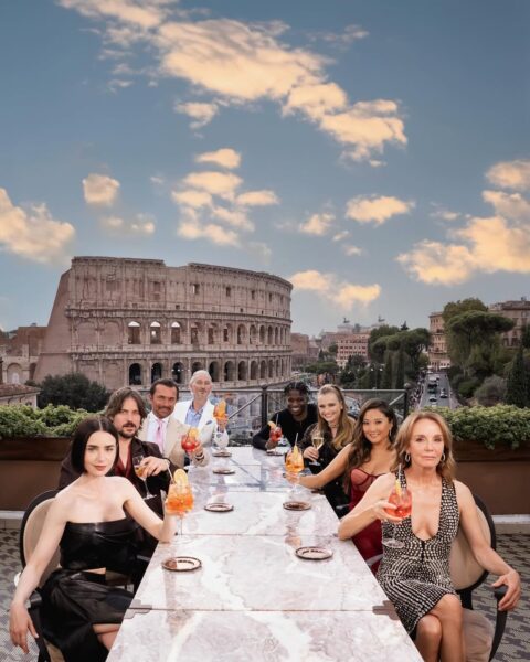 Emily i główni bohaterowie serialu siedzą przy długim stole z drinkami, na tarasie z widokiem na Koloseum i zachodzące słońce, celebrując wspólny wieczór w ikonicznej scenerii Wiecznego Miasta. Emily w Paryżu 5.