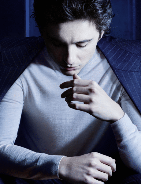 Timothée Chalamet w kampanii Bleu de Chanel, sfotografowany w półmroku z opuszczoną głową i skupionym wyrazem twarzy. Aktor ma na sobie jasny sweter i narzuconą na ramiona marynarkę w granatowe prążki, co nadaje mu tajemniczego, introspektywnego charakteru.