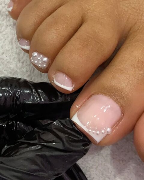 Zdjęcie stóp ze zbliżeniem na paznokcie. Są pomalowane w stylu french manicure i ozdobione perełkami.