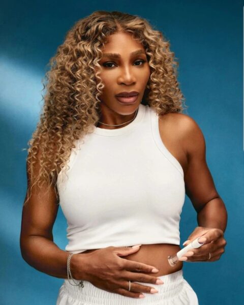 Na zdjęciu jest Serena Williams, ubrana w biały top i białe spodnie, wstrzykująca sobie lek Ozempic w brzuch. Pozuje na niebieskim tle.