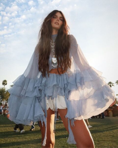 Zdjęcie influencerki, która pokazuje, jak ubrać się na festiwal 2025: ma niebieską bluzkę z falbankami w stylu boho, szorty białe oraz masywny, srebrny naszyjnik z motywem muszli. Pozuje na festiwalu. Ma długie, ciemne włosy rozpuszczone na ramiona.