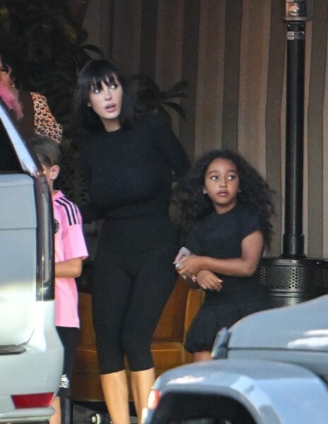 Bianca Censori dzieci: Kobieta w klasycznej czarnej stylizacji stoi obok trójki dzieci Kanye Westa i Kim Kardashian – w tym obok córki Chicago West, ubrane również w czerń. Chicago trzyma Biankę za rękę, a w tle widoczny jest chłopiec w różowej koszulce piłkarskiej. Scena rozgrywa się przy zaparkowanym SUV-ie, prawdopodobnie tuż przed wspólnym wyjazdem. Stylizacja Bianki, z prostą grzywką i dopasowanym krojem, kontrastuje z wcześniejszymi medialnymi wystąpieniami, podkreślając nowy, stonowany kierunek jej wizerunku.