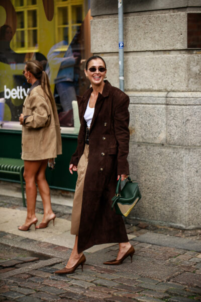 Kopenhaga trendy jesień 2025: Kobieta w eleganckim, długim płaszczu w odcieniu ciemnego brązu z podwójnym rzędami guzików, zestawionym z beżową spódnicą, białym topem i brązowymi szpilkami, niosąca zielono-beżową torebkę, uchwycona w ruchu na ulicy Kopenhagi.
