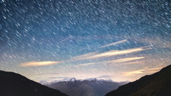 Fotografia nocnego nieba z efektem smug gwiazd w wyniku długiego czasu naświetlania, nad pasmem gór i chmurami.