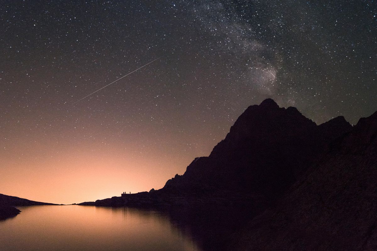 Rozgwieżdżone nocne niebo z Drogą Mleczną i spadającą gwiazdą nad górskim jeziorem, z pomarańczową poświatą na horyzoncie.