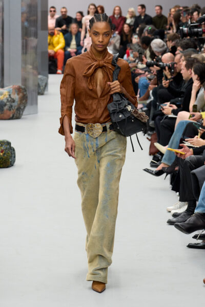Acne Studios jesień/zima 2025 – modelka na wybiegu w brązowej, wiązanej koszuli z głębokim dekoltem, szerokich przetartych jeansach z efektem sprania i skórzanych butach na obcasie. Stylizacja uzupełniona dużym pasem z ozdobną klamrą i czarną torbą na ramię, utrzymana w klimacie boho i retro.