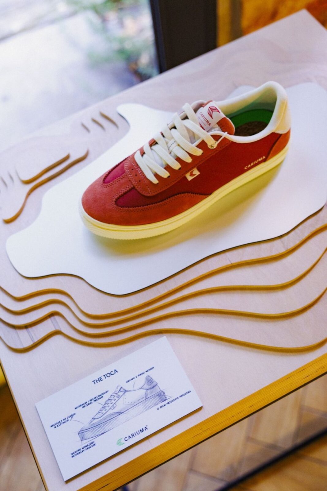 Czerwono-biały sneaker Cariuma z kolekcji The Toca zaprezentowany podczas premiery w Poznaniu, ustawiony na designerskiej ekspozycji z warstwowego drewna, symbolizującej organiczne inspiracje marki.