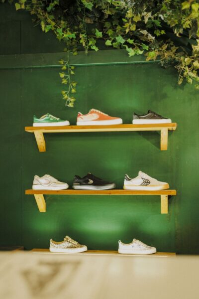 Sneakersy Cariuma w różnych modelach i kolorach, ustawione na drewnianych półkach na tle zielonej ściany z roślinnymi dekoracjami. Ekspozycja podkreśla ekologiczne DNA marki.