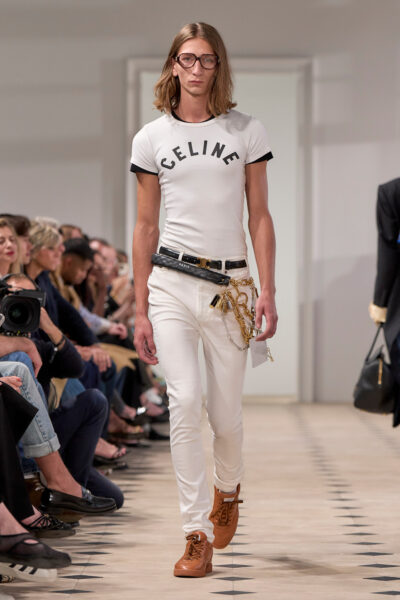 Celine wiosna/lato 2026 – model w białym dopasowanym T-shircie z logo CELINE, wąskich białych jeansach i skórzanych sneakersach w kolorze karmelowym. Stylizacja uzupełniona dużym łańcuchem z zawieszkami przy pasku i dużymi okularami o retro sznycie.