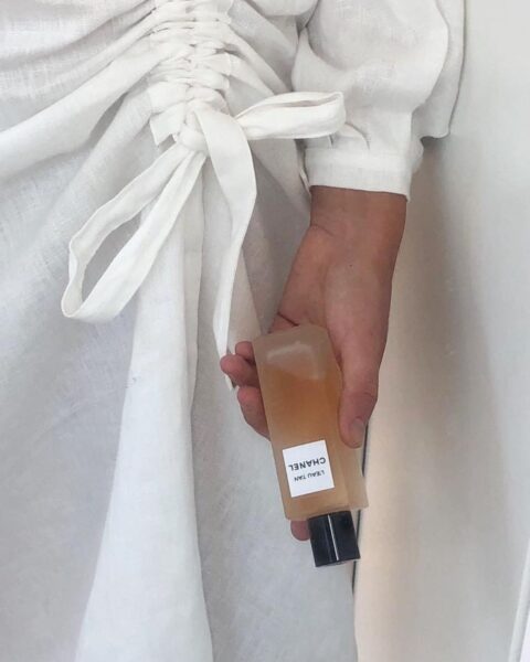 Ręka w białej lnianej sukience z marszczeniem i wiązaniem trzyma prostokątny flakon CHANEL L'EAU TAN. Subtelna elegancja w stylu premium, idealna jako wizualne odniesienie do hasła francuskie marki perfum i letniej pielęgnacji.