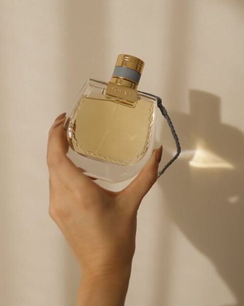 Elegancka dłoń w jasnej, zwiewnej bluzce trzyma flakon perfum Chloé Nomade Lumière d’Égypte. Bursztynowy płyn i złota zatyczka odbijają ciepłe, miękkie światło padające na jasną ścianę, tworząc cienie. Ujęcie idealnie oddaje atmosferę luksusowego, wiosennego zapachu.
