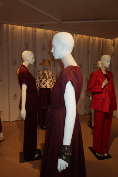 Wnętrze showroomu Max Mara, kilka manekinów ubranych w stylizacje w odcieniach burgundu i czerwieni. Najbliżej kadru sylwetka w bordowej sukience z szeroką bransoletą, w tle czerwony garnitur i stylizacja w kapeluszu.