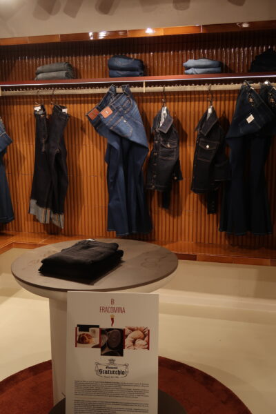 Stojak z jeansami Fracomina – elegancka ekspozycja kolekcji denim włoskiej marki Fracomina, obejmująca ciemne i klasyczne niebieskie jeansy oraz kurtki z denimu, prezentowane na tle ciepłych, drewnianych paneli. Na centralnym stole spoczywa złożona para czarnych spodni, obok tablicy informacyjnej o kolaboracji marki z historyczną cukiernią Giovanni Scaturchio z Neapolu.