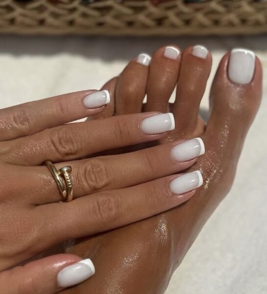 Zbliżenie na dłonie i stopy ozdobione klasycznym białym frenchem w wersji kwadratowej. Paznokcie błyszczą w świetle, a całość podkreślają złote pierścionki noszone na palcach dłoni i stóp. Stylizacja elegancka, ponadczasowa, idealna na wakacje lub specjalne okazje.