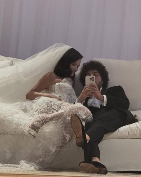 Zdjęcie za kulis ślubu w stylu vintage. Panna Młoda (Selena Gomez) w koronkowej sukni ślubnej i długim welonie siedzi obok Pana Młodego (Benny'ego Blanco) na białej sofie. Mężczyzna, ubrany w smoking, robi sobie parze zdjęcie telefonem komórkowym.