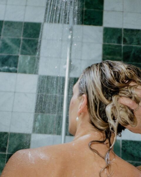 Zdjęcie kobiety stojącej pod prysznicem, która wie, jak dbać o włosy w okresie jesienno-zimowym. Myje włosy pod deszczownicą. Prysznic ma zielone kafelki.