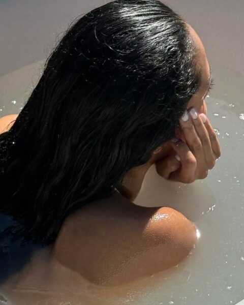 Zdjęcie kobiety leżącej w wannie, podpiera głowę o dłonie. Ma ciemne, długie włosy mokre od wody, gładko leżące na jej plecach.