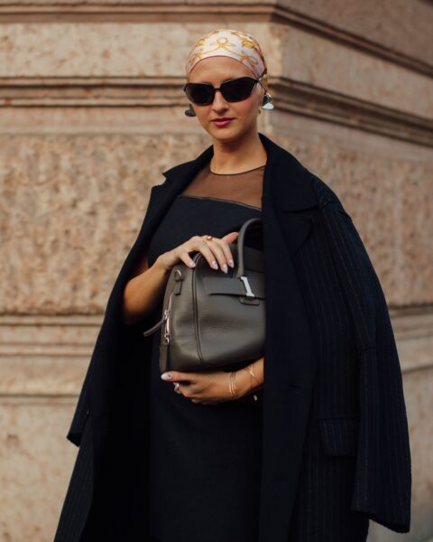 Na zdjęciu widzimy kobietę ubraną w czarną, elegancką sukienkę i ciemny płaszcz w prążki. Na głowie ma jedwabną apaszkę z modnym wzorem. Uzupełnieniem stylizacji są ciemne okulary i szaro-brązowa torebka.