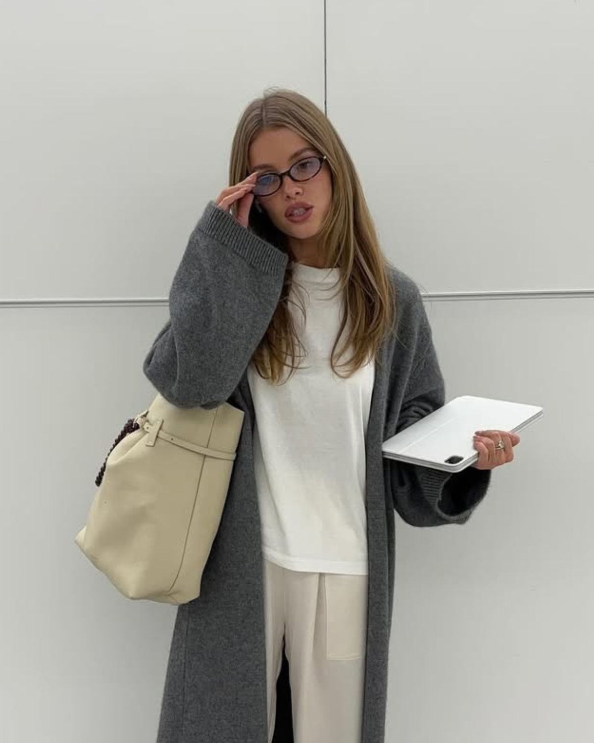 Młoda kobieta w stylowym, wygodnym stroju podróżnym. Ma na sobie długi, szary kardigan, biały T-shirt i szerokie, satynowe spodnie w kremowym kolorze. W ręku trzyma beżową torbę typu tote bag oraz laptopa. Całość wygląda bardzo elegancko, ale jednocześnie komfortowo, co idealnie wpisuje się w ideę jak stworzyć garderobę kapsułową dla studenta — stawiając na uniwersalne, łatwe do łączenia ze sobą elementy.
