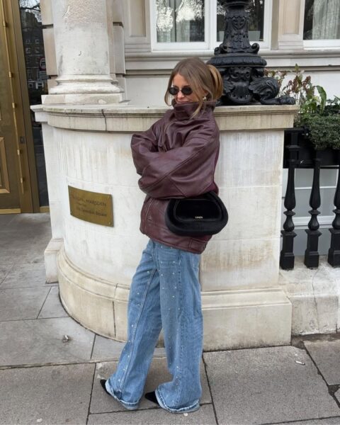 Młoda kobieta w stylowej, casualowej stylizacji na ulicy. Ma na sobie brązową skórzaną kurtkę z wysokim kołnierzem, luźne jeansy ozdobione koralikami lub małymi kryształkami, czarną futrzaną torebkę oraz czarne, prostokątne okulary przeciwsłoneczne. Całość tworzy modny i swobodny look.