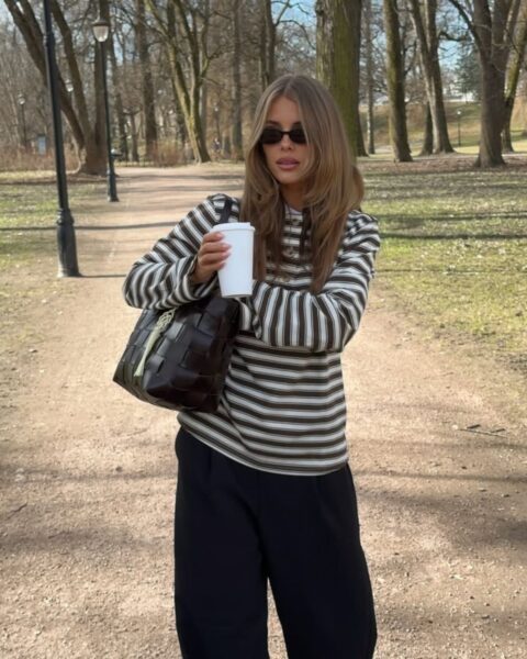 Młoda kobieta w swobodnej, jesiennej stylizacji spacerująca po parku. Ma na sobie pasiasty sweter w brązowo-białe paski, czarne spodnie z szeroką nogawką i czarne okulary przeciwsłoneczne. Na ramieniu wisi ciemna, pleciona torebka, a w dłoni trzyma biały kubek z napojem. Całość tworzy stylowy, a jednocześnie bardzo wygodny look idealny na chłodniejsze dni.
