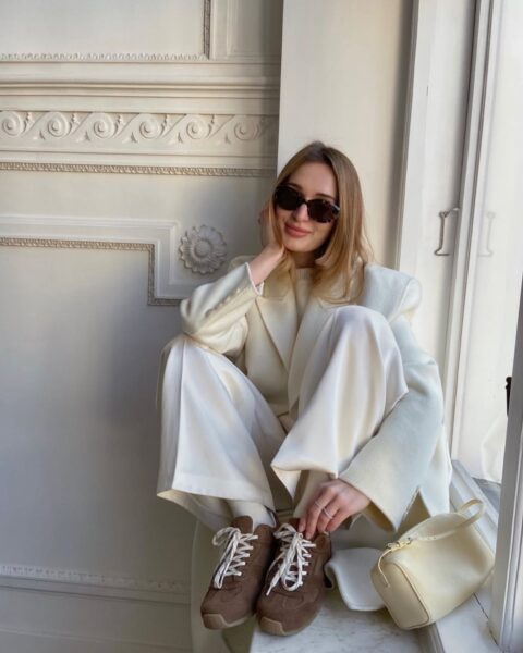 Młoda kobieta w eleganckiej, monochromatycznej stylizacji siedząca na parapecie. Ma na sobie biały płaszcz, białe szerokie spodnie i bluzkę w tym samym kolorze. Na nogach ma brązowe, zamszowe sneakersy. Obok niej leży mała, kremowa torebka. Całość stanowi udane połączenie elegancji z nutą sportowego luzu.