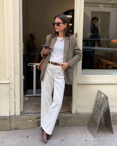 Młoda kobieta w stylowej, jesiennej stylizacji opierająca się o futrynę drzwi kawiarni. Ma na sobie tweedową marynarkę w brązowo-szarą kratę, biały T-shirt, szerokie białe spodnie z wysokim stanem, brązowy skórzany pasek i brązowe botki. Na ramieniu ma zamszową torebkę, a w dłoni trzyma telefon. Całość tworzy elegancki, a zarazem luźny i jesienny look.