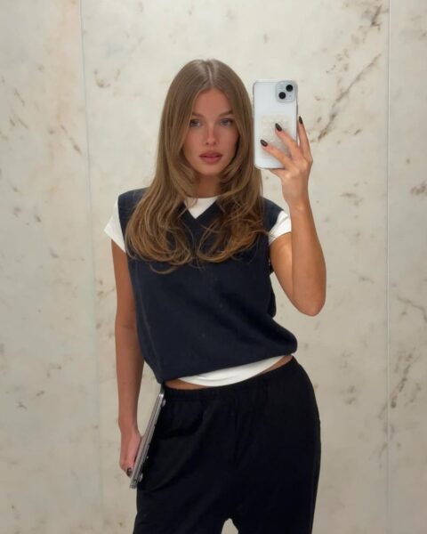 Młoda kobieta robiąca sobie selfie w lustrze, ubrana w swobodną stylizację. Ma na sobie biały T-shirt, granatową kamizelkę dzianinową i czarne spodnie dresowe. Trzyma w ręku telefon i laptopa. Całość tworzy wygodny, ale jednocześnie stylowy strój do pracy lub nauki z domu.