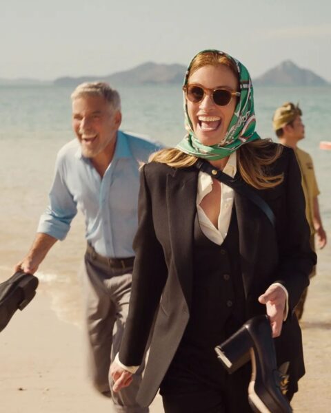 To ujęcie z komedii romantycznej "Bilet do raju" (Ticket to Paradise), którą trzeba obejrzeć, przedstawia główną bohaterkę (w tej roli Julia Roberts) w eleganckim, czarnym garniturze z kamizelką, noszącą stylowe okulary przeciwsłoneczne i zieloną apaszkę z deseniem. Kobieta idzie po piaszczystej plaży, niosąc w rękach eleganckie, czarne szpilki na słupku i uśmiecha się promiennie, patrząc wprost w obiektyw. W tle widać plażę, morze oraz George'a Clooneya, który podąża za nią, również uśmiechając się szeroko.
