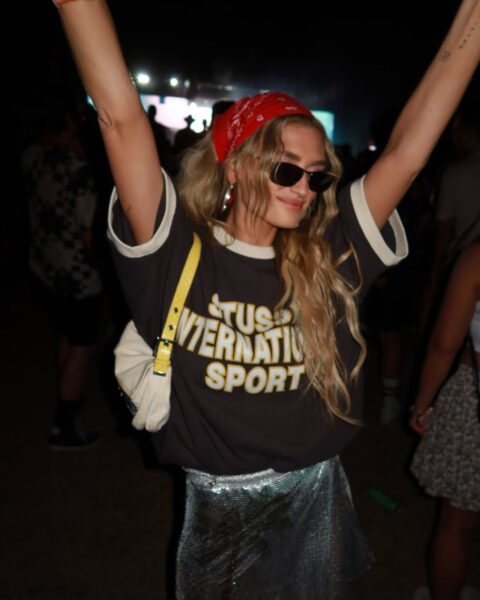 Uśmiechnięta blondynka tańczy na koncercie, z podniesionymi rękami. Ma na sobie czarny T-shirt z jasnym napisem, srebrną, błyszczącą spódnicę, okulary przeciwsłoneczne i czerwoną bandanę we włosach.