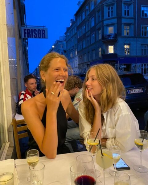 Dwie uśmiechnięte kobiety siedzą przy stoliku w letniej restauracji na zewnątrz. Mają przed sobą kieliszki z winem i drinki. Jedna z nich śmieje się głośno, druga patrzy na nią z uśmiechem. W tle widać miejską ulicę z budynkami.