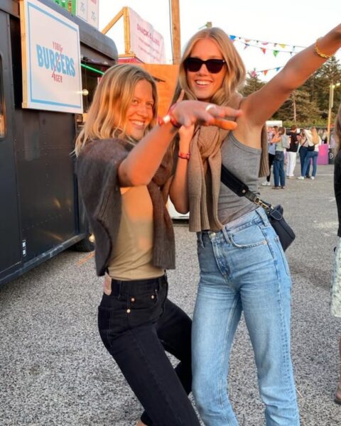 Dwie uśmiechnięte kobiety w swobodnych, festiwalowych stylizacjach tańczą i pozują do zdjęcia. Jedna z nich ma na sobie czarne dżinsy, druga niebieskie. W tle widać budkę z jedzeniem i ludzi.