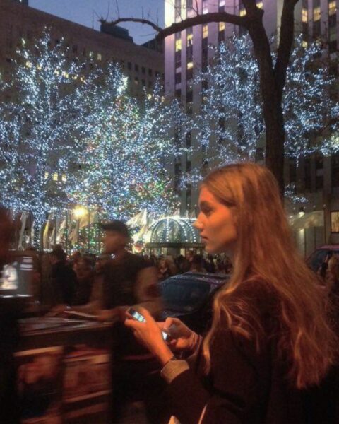 Kobieta wpatruje się w telefon, idąc ruchliwą, świątecznie oświetloną ulicą, z rozmytymi postaciami innych ludzi w tle. W oddali widać choinki ozdobione jasnymi lampkami. Scena ma klimatyczny, miejski charakter, pasujący do nocnego życia i kultury klubowej w Polsce.