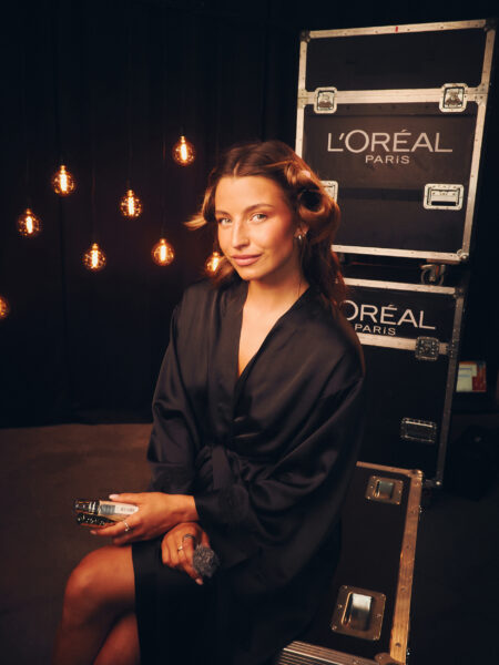 Modelka za kulisami w szlafroku podczas przygotowań do pokazu L’Oréal Paris „Pokaz Wartości” – światła sceniczne i skrzynie techniczne z logo marki w tle, stylizacja w trakcie tworzenia.