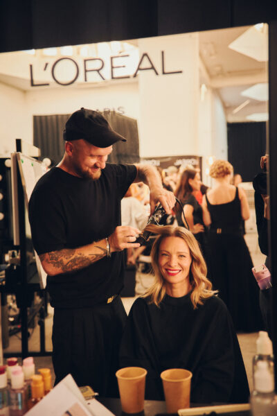 Stylista fryzur pracuje nad falami na włosach modelki w garderobie pokazu L’Oréal Paris „Pokaz Wartości”, oboje uśmiechnięci, w tle gwar przygotowań i zespół beauty.