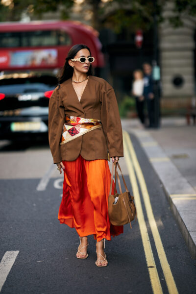 Street style z Londynu: kobieta w ekstrawaganckiej, warstwowej stylizacji – metaliczne spodnie typu cargo zestawione z futrzaną kurtką w pastelowym odcieniu i futurystycznymi sneakersami. Look uzupełnia mała torba w srebrnym kolorze i efektowne okulary przeciwsłoneczne, które nadają całości nowoczesny, kosmopolityczny charakter.