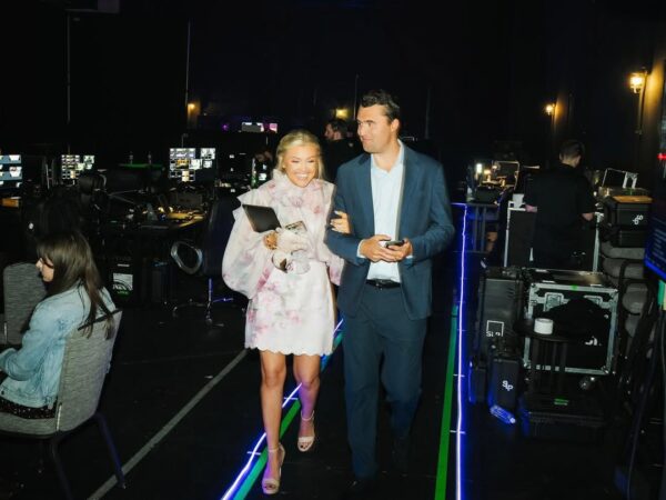 Elegancko ubrana para idzie ramię w ramię za kulisami sali z ekranami i sprzętem technicznym.