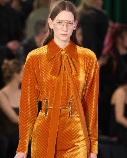 Modelka na wybiegu prezentuje stylizację z kolekcji Gucci. Ma na sobie musztardową sukienkę z aksamitu z wzorem w jodełkę. Sukienka ma duży kołnierz z wiązaniem w kokardę oraz metalowy pasek na wysokości talii. W dłoni trzyma małą torebkę z czarnej lakierowanej skóry. Modelka ma na sobie cienkie okulary w metalowych oprawkach, a jej włosy są luźno rozpuszczone.