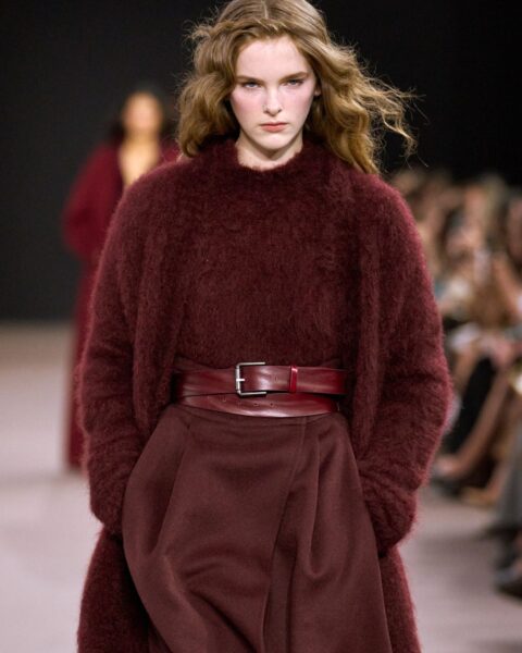 Modelka na wybiegu prezentuje stylizację z kolekcji Max Mara. Ma na sobie ciepły, puszysty sweter z moheru w kolorze bordo, który jest połączony z szerokimi, materiałowymi spodniami w tym samym odcieniu. W talii ma szeroki, skórzany pasek z prostokątną klamrą. Włosy modelki są pofalowane i sprawiają wrażenie lekko potarganych. Tło jest rozmyte, widać na nim sylwetki innych osób.