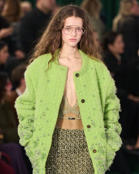 Modelka prezentuje stylizację z kolekcji Gucci na wybiegu. Ma na sobie limonkowe, futerkowe bolerko z ozdobnymi, błyszczącymi aplikacjami, pod którym widać delikatny biustonosz w beżowym kolorze. Do tego nosi spódnicę ołówkową z czarno-złotym, tweedowym wzorem. Na nosie ma okulary w cienkich, metalowych oprawkach, a jej włosy są luźno rozpuszczone.
