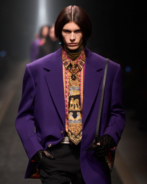 Model prezentuje na wybiegu stylizację z kolekcji Versace. Ma na sobie czarny krawat w orientalne wzory, purpurową marynarkę z poduszkami na ramionach oraz czarne spodnie. W dłoni trzyma małą czarną torebkę. Tło zdjęcia jest ciemne, widać na nim sylwetki innych osób. Całość to przykład na najbardziej zaskakujące kolory mody 2025: połączenie fioletu z żółcią i wzorami.