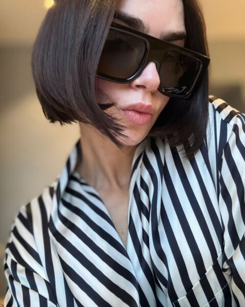 Zdjęcie Lily Collins, która ma ciemne włosy w stylu jawline cut. Włosy są proste, ale podwinięte do wewnątrz na końcach. Na nosie ma okulary przeciwsłoneczne. Ubrana jest w koszulę w czarno-białe paski.