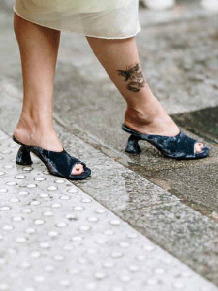 Buty typu mule w granatowym welurze, na nietypowym, szerokim obcasie, zestawione z jasną spódnicą midi. Stylizacja sfotografowana podczas deszczu, z widoczną mokrą nawierzchnią chodnika i tatuażem na łydce.
