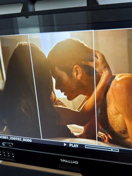 Backstage z planu filmowego – kamera uchwyciła monitor podglądowy, na którym widać intymną scenę pocałunku kobiety i mężczyzny w ciepłym, złotym świetle poranka. Obraz ukazuje atmosferę bliskości i sensualności, a techniczne ramki ekranu nadają ujęciu surowy, filmowy charakter.