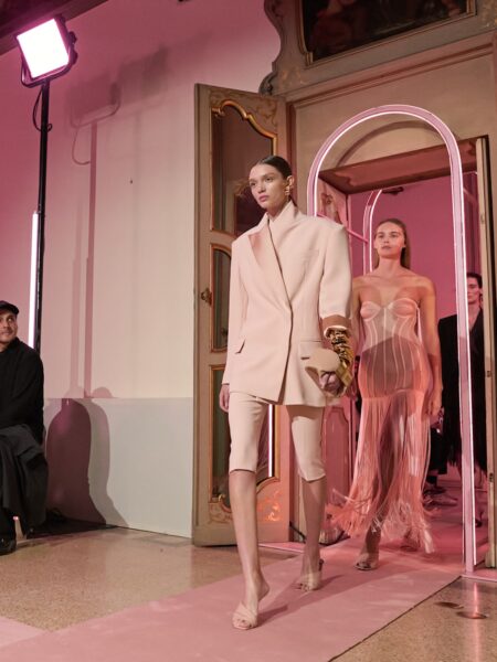 Milan Fashion Week 2026: Modelki wchodzące na wybieg w bogato zdobionej sali z różowym światłem i drewnianymi zdobieniami. Pierwsza prezentuje garnitur w kolorze pudrowego beżu z krótkimi spodniami, kolejna ma na sobie przezroczystą, tiulową suknię z gorsetową górą. Publiczność uważnie obserwuje kolekcję.